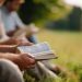 young group reading and discussing bible outdoors, christian fellowship and prayer meeting at park, outdoor scripture study, nature worship gathering, faceless participants, defocused